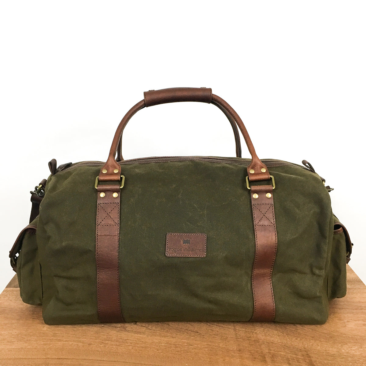 A Rogue Industries White Cap Waxed Canvas Duffle with full-grain leather straps and handles is placed on a wooden surface against a plain white background.